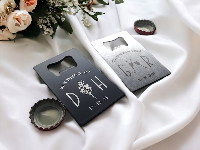 May include: Two rectangular bottle openers with a bottle cap. One is dark gray with white text and a floral design, the other is silver with text and champagne glasses. The dark gray opener says "SAN DIEGO, CA" and "D H 12.12.24". The silver opener says "Cheers to many years! GAR 04-24-2024".