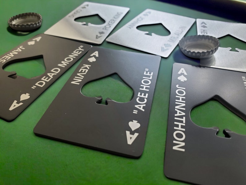 May include: A collection of rectangular metal bottle openers shaped like playing cards, featuring spade-shaped cutouts and custom text including names like James, Kevin, and Johnathon, alongside phrases like "Dead Money" and "Ace Hole," with two bottle caps.