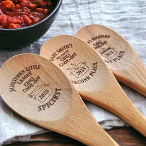 May include: Three wooden spoons with engraved text and a chili pot graphic. The spoons are labeled for a chili cook-off, with text including "Jamestown Little League Chili Cook-Off 2023 Spiciest", "Gary Smith's 3rd Annual Chili Cook-Off 2023 Second Place", and "N's Backyard Chili Cook-Off 2023 1st Place".