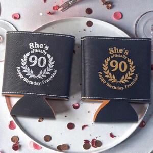 May include: Two black leatherette can coolers featuring white stitching and a circular laurel wreath design. The text reads "She's officially 90" and "Happy Birthday, Francine!" One cooler has white lettering, while the other features gold-colored lettering.