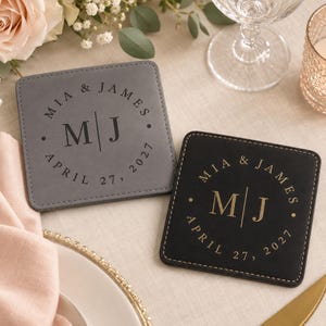 May include: Two square coasters, one gray and one black, featuring stitched edges. Each coaster is personalized with the names MIA & JAMES, the initials M | J, and the date APRIL 27, 2027, printed in a circular layout on the surface.