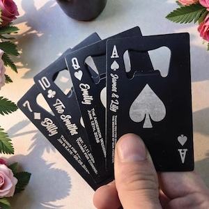 May include: A set of four black, rectangular bottle openers shaped like playing cards. Each opener features a suit symbol, a rank, and custom engraved text including names and dates such as Billy, The Smiths, Emily, and James & Lily.