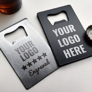 May include: Two rectangular bottle openers, one silver and one black, with the text "YOUR LOGO HERE" in white. The silver opener has five stars and the word "Engraved". A bottle cap is next to the black opener.