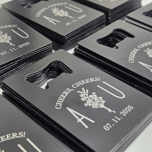 May include: Black bottle openers with a rectangular shape and a built-in bottle opener. Each opener is engraved with the words "Cheers, Cheers!" and the initials "A U" along with the date "07.11.2026" and a floral design.