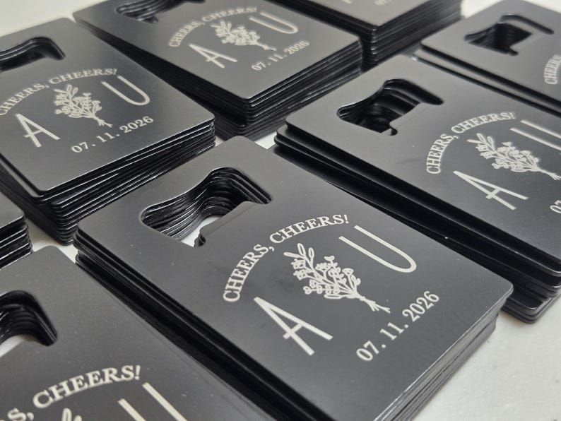 May include: Black bottle openers with a rectangular shape and a built-in bottle opener. Each opener is engraved with the words "Cheers, Cheers!" and the initials "A U" along with the date "07.11.2026" and a floral design.