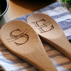 May include: Two wooden spoons with engraved initials and names. One spoon has the letter "S" with "SHARP" engraved, and the other has the letter "E" with "ELLIS" engraved. The spoons are light brown and rest on a striped cloth.