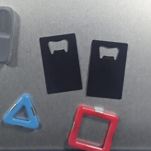 May include: An assortment of bar accessories and magnets, including two black bottle openers, a blue triangle, a red square, and a gray rectangular magnet. The items are arranged on a metallic surface.