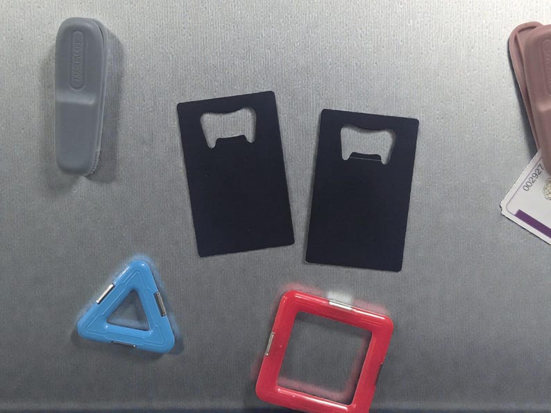 May include: An assortment of bar accessories and magnets, including two black bottle openers, a blue triangle, a red square, and a gray rectangular magnet. The items are arranged on a metallic surface.