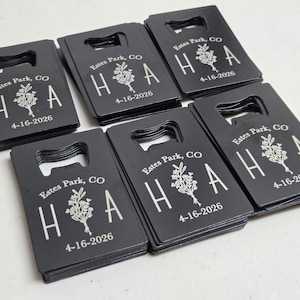 May include: A collection of black bottle openers with a rectangular shape and a built-in bottle opener. Each opener is engraved with the text "Estes Park, CO", the letters "H A", a floral design, and the date "4-16-2026" in white.