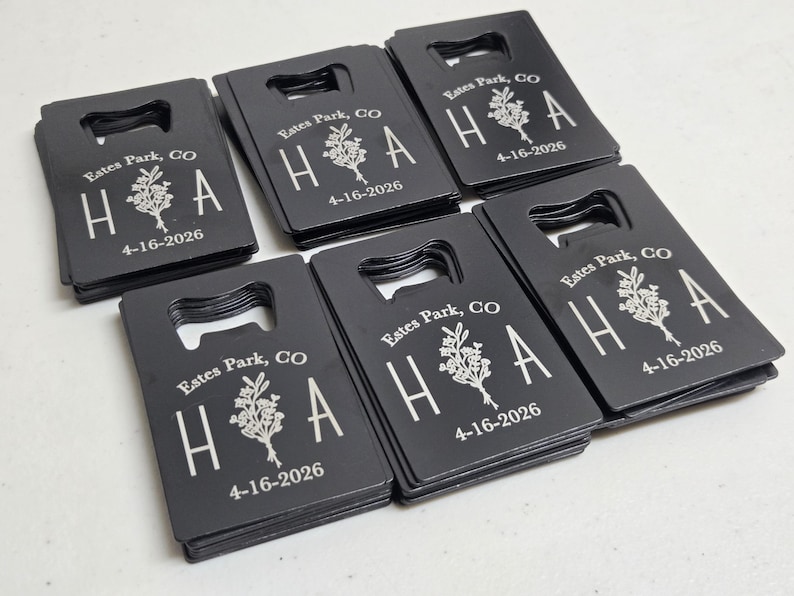 May include: A collection of black bottle openers with a rectangular shape and a built-in bottle opener. Each opener is engraved with the text "Estes Park, CO", the letters "H A", a floral design, and the date "4-16-2026" in white.