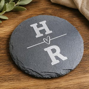 May include: A round, dark gray slate coaster featuring a rough, natural edge. The center is engraved with the letter H above a horizontal line, a small heart symbol, and the letter R below the line, all in a light gray, matte finish.