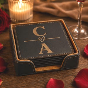 May include: A stack of square black coasters with tan stitching, housed in a matching black holder. The top coaster features a gold-toned engraved design with the letter C above a horizontal line containing a small heart, followed by the letter A.