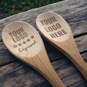 May include: Two light-colored wooden spoons featuring laser-engraved text. One spoon displays the text "YOUR LOGO HERE" above five stars and the word "Engraved". The second spoon displays the text "YOUR LOGO HERE" in a simple block font.