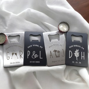 May include: Four personalized bottle openers with silver and black finishes are displayed on a white cloth. Each opener has a different design and text, including names, dates, and locations like New York and San Diego. Bottle caps are placed near the openers.