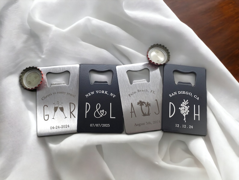 May include: Four personalized bottle openers with silver and black finishes are displayed on a white cloth. Each opener has a different design and text, including names, dates, and locations like New York and San Diego. Bottle caps are placed near the openers.