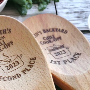 May include: Two light-colored wooden spoons with dark laser-engraved text and graphics. One spoon reads "3rd Annual Chili Cook-Off 2023 Second Place" with a pot illustration. The other reads "Chili Cook-Off 2023 1st Place" with a pot illustration.
