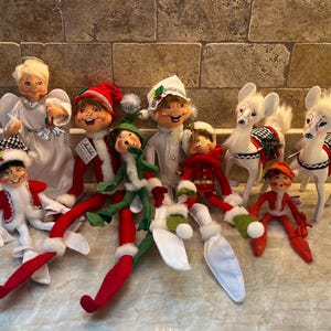 May include: A collection of vintage-style Christmas figurines including an angel in a white gown, several elves in red, green, and white outfits with pointed hats, and two white deer with faux fur accents and decorative saddles, all arranged together.