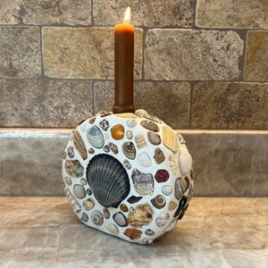 May include: A circular white candle holder decorated with a mosaic of various seashells in shades of brown, gray, and white. A single brown taper candle is inserted into the top center of the holder, with a small flame burning at the wick.
