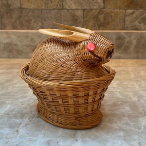 May include: A woven wicker bunny-shaped basket with a lid. The bunny has upright bamboo ears and a red bead for a nose. The basket is light brown and has a round base.