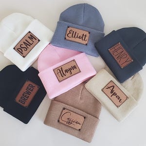 May include: A collection of seven knit beanies in various colors, including white, black, pink, and gray. Each beanie features a rectangular leather patch with a name or word, such as "Psalm" or "Elliott".
