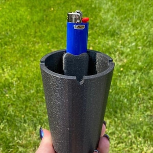 May include: A black, cylindrical ashtray with a built-in lighter holder. A blue lighter with the word "BIC" printed on it is inserted into the holder.