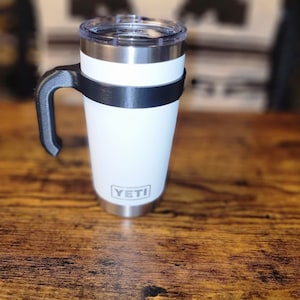 May include: White YETI travel mug with a black handle and lid. The mug is sitting on a wooden surface.