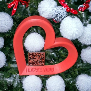 May include: A red heart-shaped ornament with the words "I LOVE YOU" and a black and white QR code. The ornament is surrounded by white fluffy balls and green pine branches.