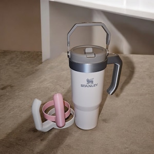 May include: A white and gray Stanley travel mug with a black logo and a gray handle. The mug is sitting on a gray surface with a pink silicone lid.