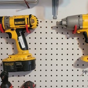 May include: Two yellow DeWalt power tools hanging on a pegboard. The tool on the left is a drill with a black battery and a red drill bit. The tool on the right is a yellow impact wrench with a gray head.