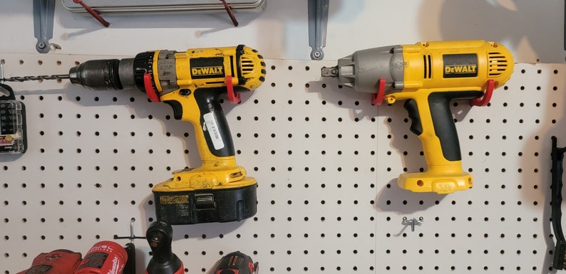 May include: Two yellow DeWalt power tools hanging on a pegboard. The tool on the left is a drill with a black battery and a red drill bit. The tool on the right is a yellow impact wrench with a gray head.
