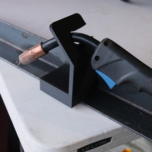 May include: A black plastic welding torch holder with a copper tip. The holder is resting on a metal surface. The torch is labeled "Miller".