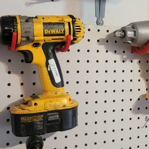 May include: Two yellow and black power tools hanging on a pegboard wall. The tool on the left is a cordless drill with a black and silver chuck. The tool on the right is a cordless impact driver with a silver head. Both tools have red accents and the word "DEWALT" printed on them.