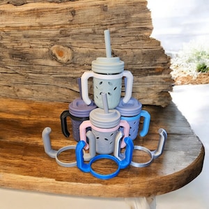 May include: A set of five silicone baby cups with handles and lids. The cups are stacked on top of each other and are in various shades of blue and pink. The cups are on a wooden surface.