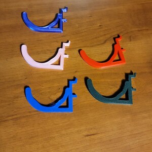 May include: Five 3D printed wall hooks in different colors: blue, pink, red, dark blue, and black. The hooks are shaped like a curved bracket with a flat base.