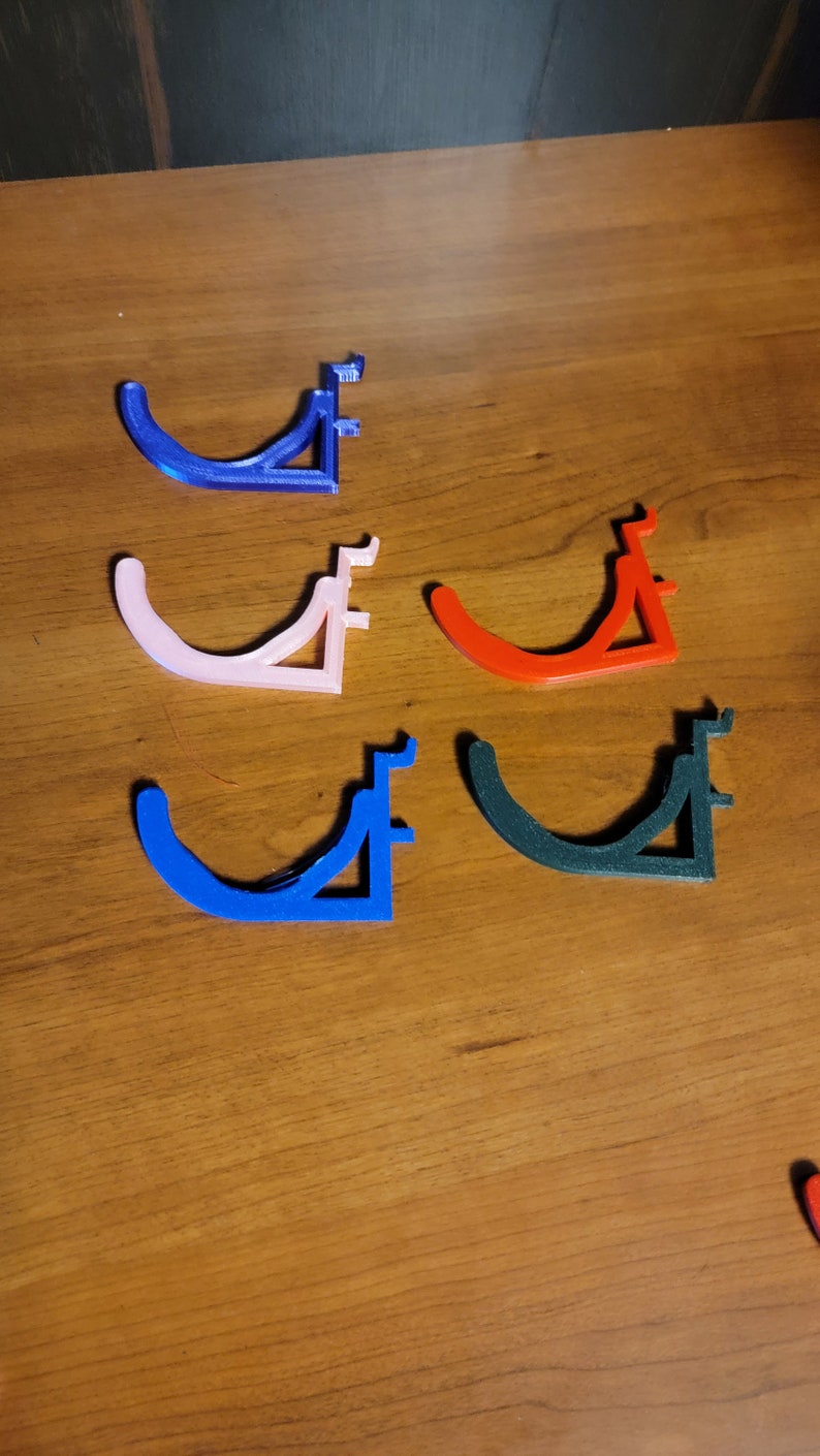 May include: Five 3D printed wall hooks in different colors: blue, pink, red, dark blue, and black. The hooks are shaped like a curved bracket with a flat base.