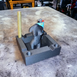 May include: A black ashtray with a female torso sculpture, a pre-rolled cigar, and a blue lighter. The ashtray has a rectangular shape with rounded corners and circular indentations for holding items.