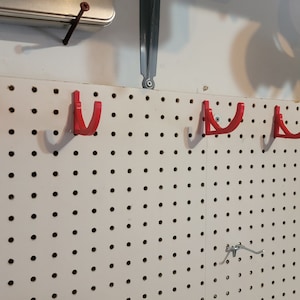 May include: A white pegboard with five red plastic hooks mounted on it. The hooks are designed to hold tools or other items. There are also some tools hanging on the pegboard.