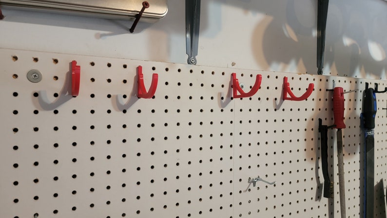 May include: A white pegboard with five red plastic hooks mounted on it. The hooks are designed to hold tools or other items. There are also some tools hanging on the pegboard.