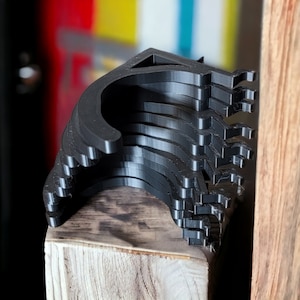 May include: A stack of black plastic door stops, each with a curved shape and a hook on the end. The door stops are arranged in a pile on a wooden surface.