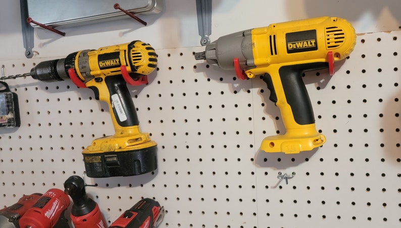 May include: Two yellow DeWalt power tools hanging on a pegboard wall. The tool on the left is a cordless drill with a black battery. The tool on the right is a cordless impact driver with a gray head.