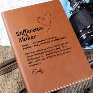May include: A brown leather journal with the words "Difference Maker" and a heart graphic. The journal includes a definition and a personalized name, "Emily". The journal is on a map and a travel bag.