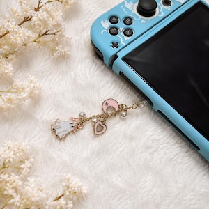 May include: A light blue handheld gaming console with a gold-toned metal charm attached to the side. The charm features a white ghost, a pink crescent moon with a star, and a pink heart, all accented with small white faux pearls on a gold chain.