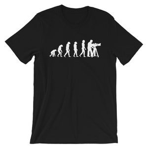 May include: Black t-shirt with a white graphic depicting the evolution of a videographer. The design shows the stages of human evolution leading to a person operating a video camera on a tripod. A fun design for video makers.