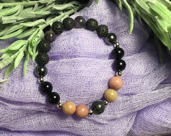 Può includere: Un braccialetto fatto di perline di roccia lavica nera, ametista, ematite, onice nero e quarzo rosa. Il braccialetto è su uno sfondo di tessuto viola.