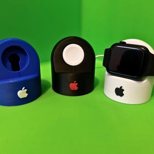 Puede incluir: Tres soportes de carga para Apple Watch impresos en 3D. Uno es azul, otro es negro y otro es blanco. Cada soporte tiene un logotipo de Apple en la parte delantera.