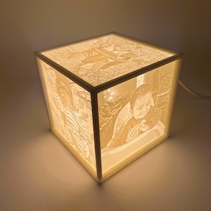 Pode incluir: Uma lâmpada branca em forma de cubo com uma imagem 3D de duas crianças dentro. A lâmpada está acesa e a imagem está iluminada.