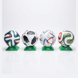Pode incluir: Quatro bolas de futebol em miniatura em suportes verdes. As bolas apresentam designs dos torneios 2014 Brazuca, 2018 Telstar 18, 2022 Al Rihla e 2026 Trionda. Cada suporte está rotulado com o ano, o país anfitrião e o nome da bola.
