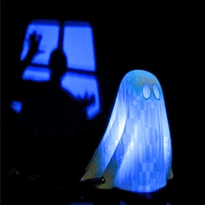 May include: A blue glowing ghost figurine with two white eyes. The ghost is sitting on a dark surface in front of a window with a blue glow.