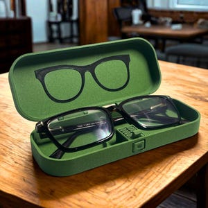 Pode incluir: Um estojo retangular para óculos em verde-oliva mate com tampa articulada. O interior apresenta um contorno preto moldado de armações de óculos na tampa e um par de óculos pretos Tom Ford repousando dentro do estojo sobre uma ponte.