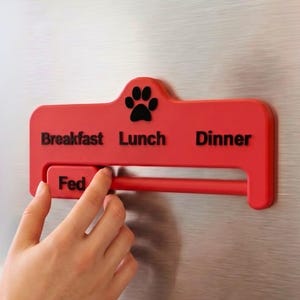 Może przedstawiać: Czerwony przypomnienie o karmieniu zwierząt domowych z nadrukowanymi słowami "Breakfast", "Lunch" i "Dinner". Odbicie łapy znajduje się nad słowem "Lunch". Słowo "Fed" znajduje się na przesuwnym pasku. Ręka przesuwa pasek.